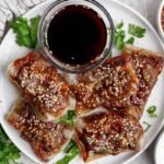 A white plate with five pieces of crispy, glazed tofu topped with sesame seeds, garnished with cilantro, and served with a small bowl of dark dipping sauce.