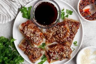 A white plate with five pieces of crispy, glazed tofu topped with sesame seeds, garnished with cilantro, and served with a small bowl of dark dipping sauce.