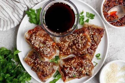 A white plate with five pieces of crispy, glazed tofu topped with sesame seeds, garnished with cilantro, and served with a small bowl of dark dipping sauce.
