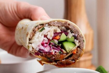 A hand holds a stuffed wrap filled with rice, vegetables, and greens over a bowl of dipping sauce, with a plate of salad in the background.