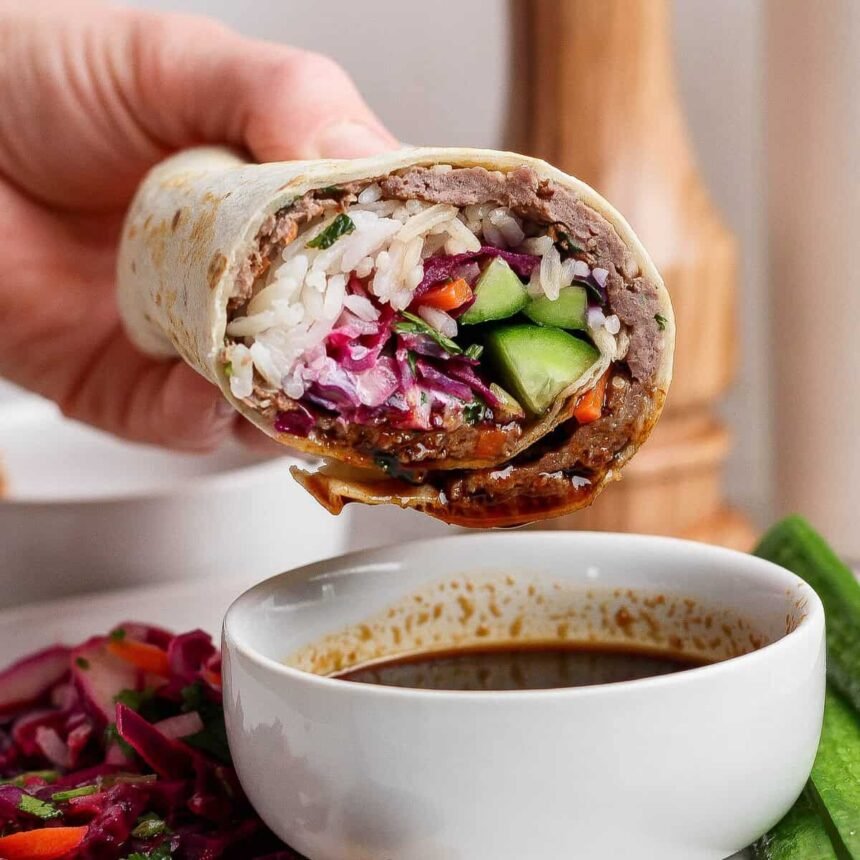A hand holds a stuffed wrap filled with rice, vegetables, and greens over a bowl of dipping sauce, with a plate of salad in the background.