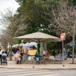 A kiosk cafe in a Lisbon garden square under bare plane trees, with people in coats and scarves sitting at metal tables and market stalls set up behind them