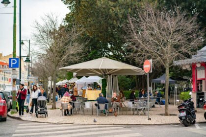 A kiosk cafe in a Lisbon garden square under bare plane trees, with people in coats and scarves sitting at metal tables and market stalls set up behind them