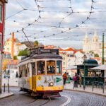 A yellow vintage Lisbon tram rounding a curve on cobblestone tracks with pastel buildings and the domed church of Sao Vicente de Fora rising in the background