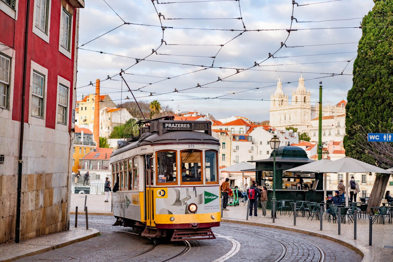A yellow vintage Lisbon tram rounding a curve on cobblestone tracks with pastel buildings and the domed church of Sao Vicente de Fora rising in the background