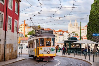 A yellow vintage Lisbon tram rounding a curve on cobblestone tracks with pastel buildings and the domed church of Sao Vicente de Fora rising in the background