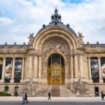 The gilded entrance gates of the Petit Palais in Paris under a flat grey overcast sky, with only a few people crossing the avenue