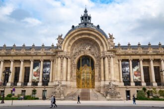 The gilded entrance gates of the Petit Palais in Paris under a flat grey overcast sky, with only a few people crossing the avenue